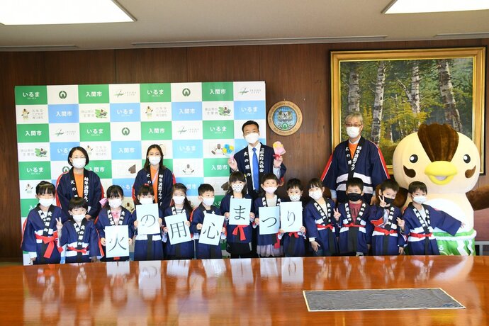 入間市と書かれたパネルと額絵の前で前列に木の実保育園の園児13名、後列左から眼鏡とマスクを着用した女性、白いマスク姿の長髪の女性、手作りの花束を両手に持った杉島市長、金髪のマスク姿の男性、入間市のマスコットキャラクターいるティーの着ぐるみを着た人を撮影した写真、いるティー以外は火の用心の法被を着ている