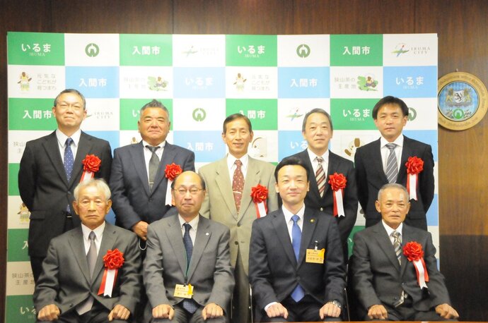 入間市技能功労者表彰式の際に入間市と書かれたパネルの前で前列に椅子に座った4人の男性、後列に起立した5名の男性撮影した写真、杉島市長は前列の右から2人め