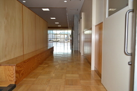 奥に日が差す大きな窓が見え、左に木製の長い椅子、右に室内への白い引き戸がある廊下を、正面から撮影した写真