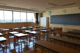 奥に日が差す複数の窓が並び、フローリングの上に黒板と複数の机と椅子、会議テーブルが並べられた教室の写真