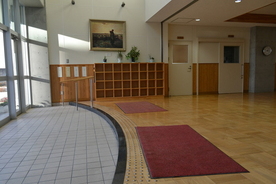 大きなアーチ状の上がり框と奥に手摺を備えた大きな玄関と、上がり框を上がった上に広がるフローリングの上に敷かれた2枚の大きな赤色のカーペットがあり、奥に2つの扉と壁に絵画が飾られている武蔵中学校を撮影した写真