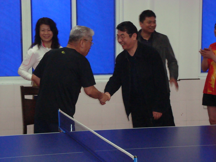 卓球台の横で、握手をする男性2名、その横で笑顔な男性と女性の写真。