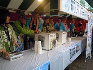 緑と白のイベントテントの下で、緑や水色の法被を着たドイツ人と日本人が、テーブルの上に設置されたビールサーバについて話している様子を撮影した写真