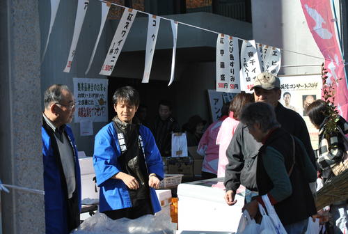 佐渡市の名物を使用した料理の名前が書かれた複数の白い紙が吊りさげられた下で青色の法被を着た男性2名と帽子をかぶった男性と眼鏡をかけた女性が会話している様子を正面から撮影した写真、奥にも5名の男女が映っている。