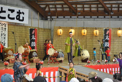 緑のハッピを着た演者がマイクの前に立っている。うしろに和太鼓の人たちを4名従えている。子供が2人舞台の下でそれを聞いている。他にも観客の頭が見える。