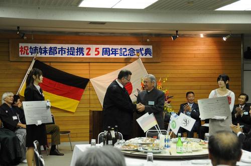 ウッド調の壁に「祝 姉妹都市提携25周年記念式典」と書かれたステージ看板とドイツ国旗が飾られ、その前に並んで座る両国の男女と、立って握手を交わす両国の男性を撮影した写真