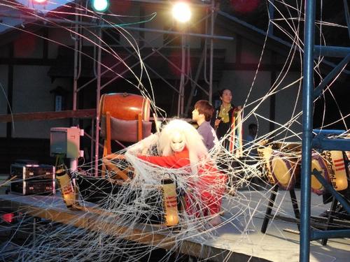 赤い着物を着た白い長髪の演者が蜘蛛の糸のようなものを舞台に放っており、後ろには和楽器の演奏者がいる