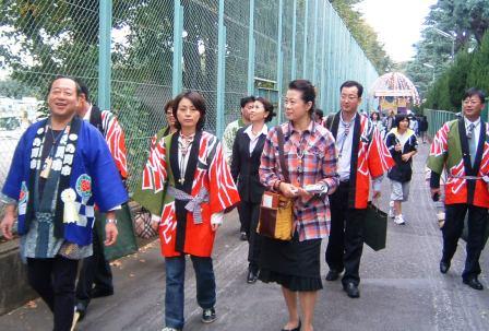 私服、スーツ、青色の法被、赤色の法被を着た複数人の男性と女性が金網の横を歩いている写真。