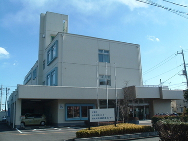 青空の下、白い壁の3階建ての男女共同参画推進センター外観を撮影した画像で、入口の横には障がい者用駐車スペース、旗ポールが3本、植木等がある。