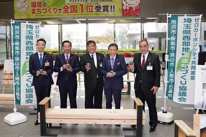 所沢市、飯能市、狭山市、入間市、日高市の市長5名がはしらベンチの後ろに立って笑っている様子を写した写真
