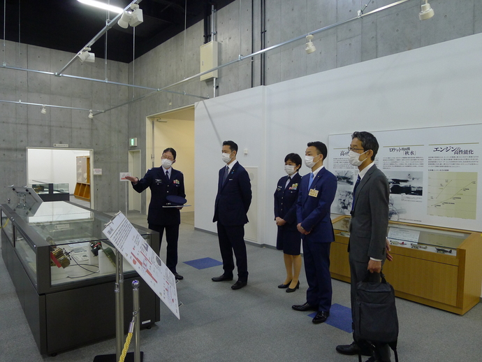 博物館の中で、スーツ姿の関係者が立って並び、職員から説明を聞いている写真