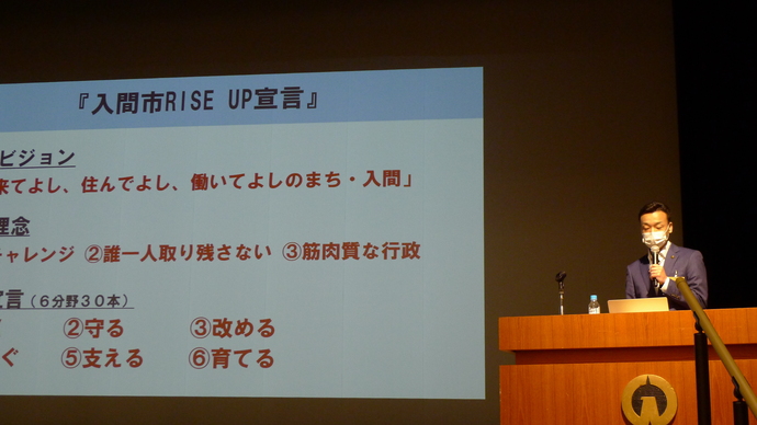 入間市RISEUP宣言と映し出されたスクリーンの右前方の壇上でマスク姿の杉島市長がマイクを左手に持って立っているところを斜め前から撮影した写真