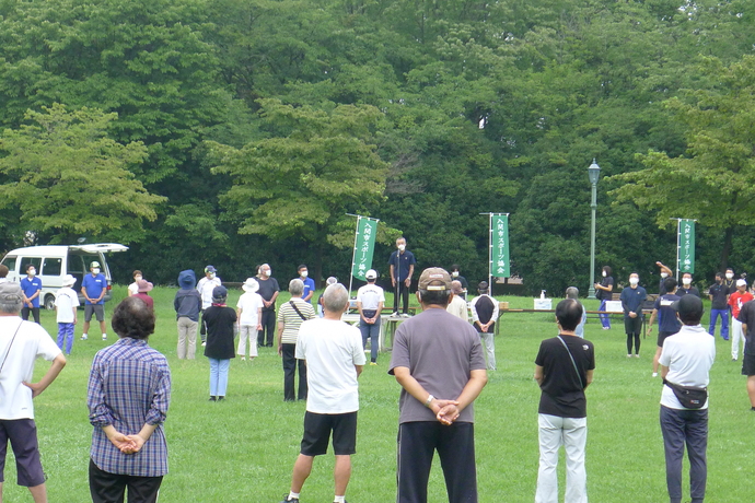 晴天の下、緑の草木が生い茂った彩の森入間公園でマイクが備え付けられた中央の台に立った男性を囲むようにラジオ体操をしているたくさんの人たちを撮影した写真、会場には入間市スポーツ協会と書かれたのぼり旗が3本立てられている