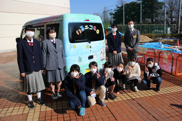 制服を着た女子学生と、私服姿の子どもたちが、イラストが描かれた水色の車の周りに集まっている集合写真