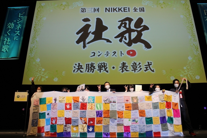 「第三回NIKKEI全国社歌コンテスト 決勝戦・表彰式」と表示されたスクリーンの下で、カラフルな横断幕を持った出演者たちが並んでポーズをとっている写真