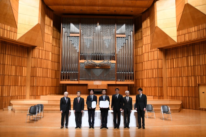 大きなパイプオルガンがある音楽ホールで、スーツ姿の男性たちが協定書を手に持って並んでいる写真