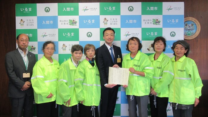明るい緑色のジャケットを着た女性たちと、スーツ姿の男性が並び、中心の二人が紙を持って立っている写真