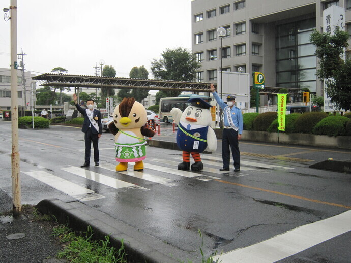 くもり空の下、横断歩道で左から杉島市長、入間市のマスコットキャラクターのいるティー、埼玉県警察のマスコットキャラクターぽっぽくん、計4名が縦一列に並んで片手をあげて歩いている様子を撮影した写真
