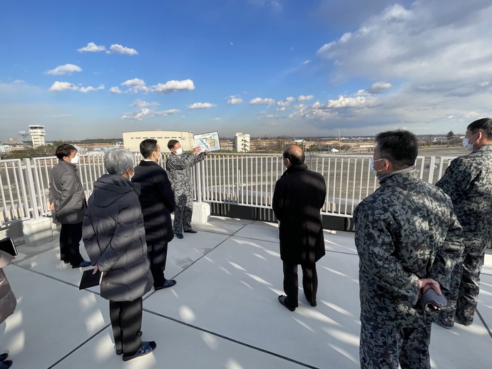 建物の屋上に上着を着た人たちが集まり、男性の説明を聞いている写真