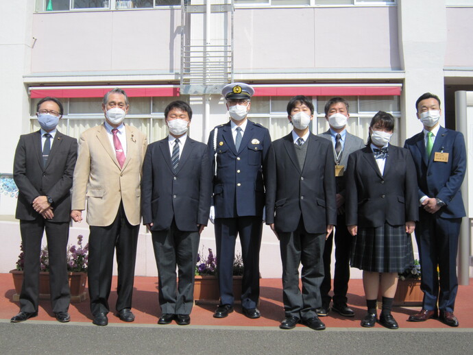 学校の建物の前で、制服姿の男性を中心に、関係者や学生たちが横に並んでいる写真