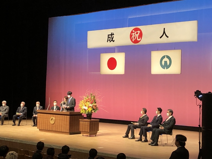 奥に祝成人と書かれた横断幕とその左下に日本国旗、右下に入間市市章が 掲げられたホールで、手前にステージを見守る観客、ステージの演壇にマスクと両手で紙を持ったスーツ姿の男性とその左脇に女性が立っており、2人の左右には計7名のマスクを付けたスーツ姿の男性が椅子に座っている入間市成人式の様子を撮影した写真