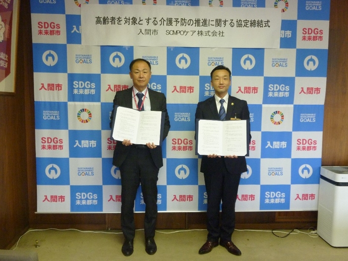 インタビューボードの前でスーツ姿の男性2人が、書類を手に持って並んでいる写真