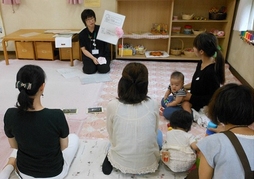 訪問時の写真。絨毯敷きの室内で、説明用の紙を持って女性が説明をしており、乳幼児を連れた親子4組がその話を聞いている。
