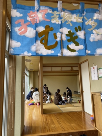 「子育て支援センターむぎ」と書かれた青空模様の暖簾があり、その向こうの和室で親子が遊んでいる写真。