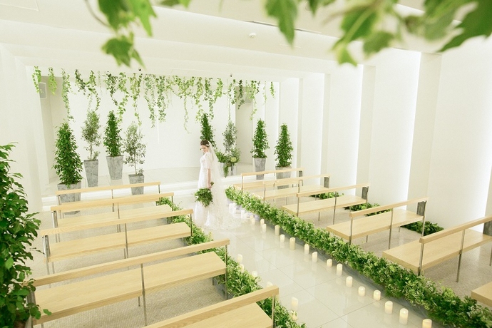 長椅子のそばにキャンドルと観葉植物が複数置かれた式場にブーケを持った花嫁が立っている写真