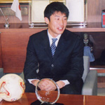 サッカーボールの乗っている机にスーツ姿の男性が座っている写真