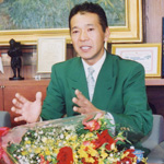 花束の置かれた机の前で緑のスーツ姿の男性が手を動かしながら話している写真