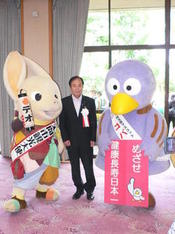 スーツを着た男性がタスキをかけたマスコットキャラクター2体の間に立っている写真