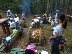 林の中にあるキャンプ場での焚火の様子。児童らが3名程度で班をつくり、大人たちが見守るなかで、3つの班がそれぞれ焚火を囲んでいる写真。