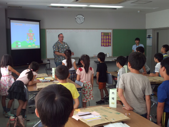 スクリーンに絵が映されていてその横に外国人の男性がおり、手前の子どもたちが立っている写真