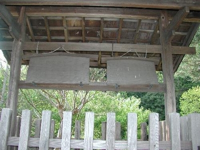 木の柵の奥に、木の屋根の柱から下がる2つの大きな絵馬型の木看板の写真