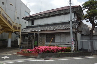 二階建てで瓦屋根の古い日本家屋の前に咲くつつじと電話ボックスがある写真