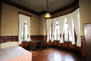 白いカーテンの大きな窓と天井から下がるランプとベッドの置かれた部屋の写真