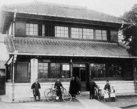 二階建て瓦屋根の日本家屋の前に立つ人たちと自転車のモノクロ写真